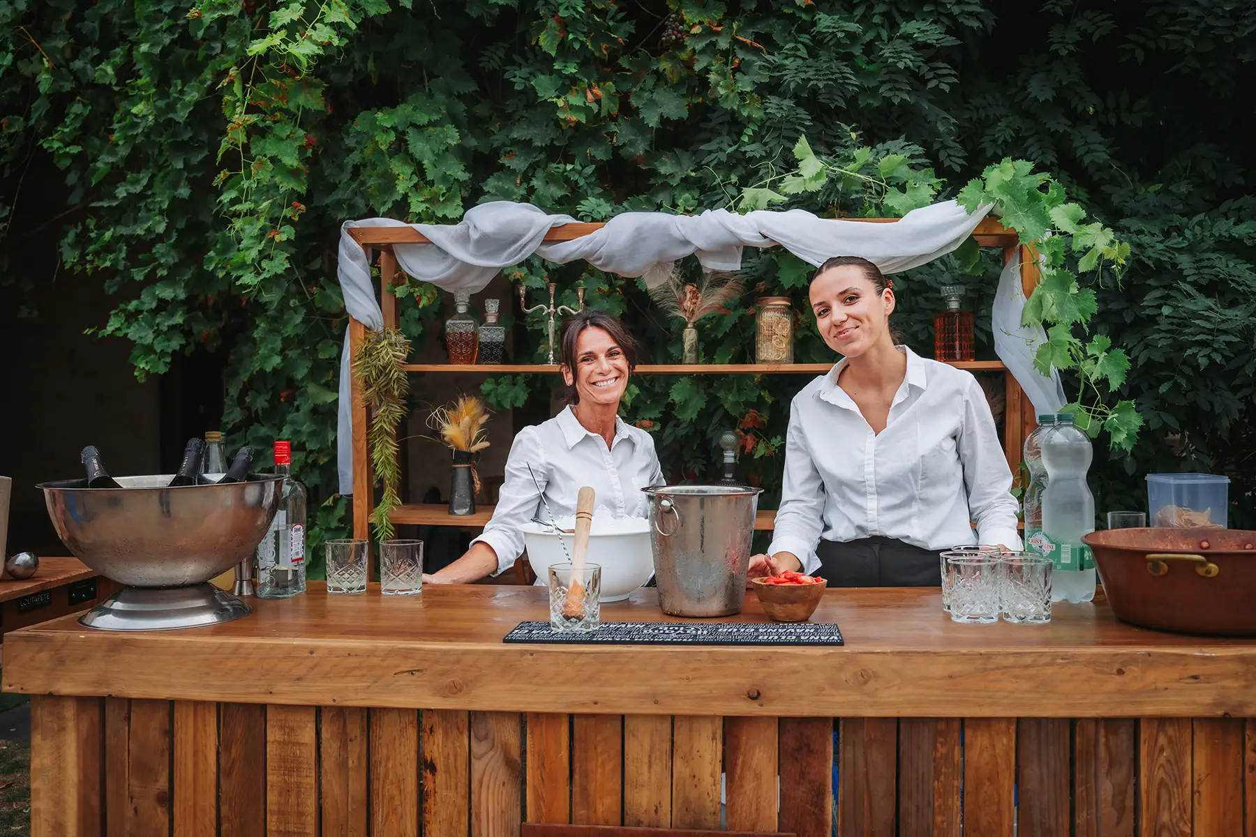 Aurélie & Jérémy 99 Deux barmaids souriantes posant derrière un bar en bois décoré en extérieur