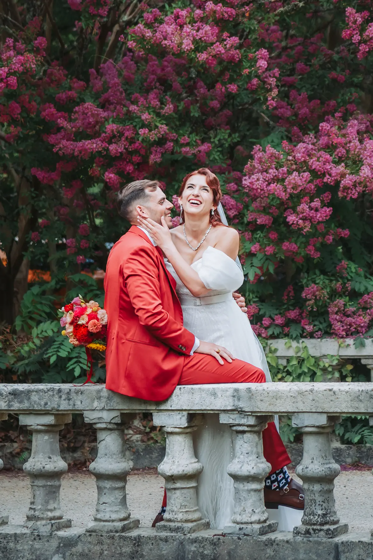 Aurélie & Jérémy 96 Le marié en costume rouge et la mariée riant assis sur une balustrade en pierre